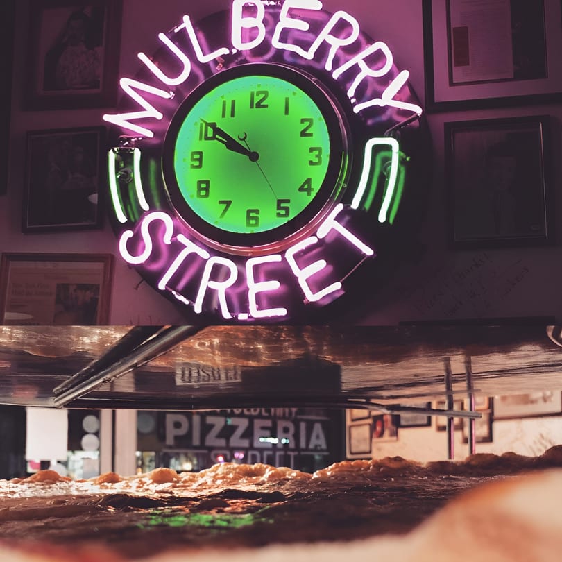 A neon "Mulberry Street" clock glows above a pizza, with a "Pizzeria" sign in the background.