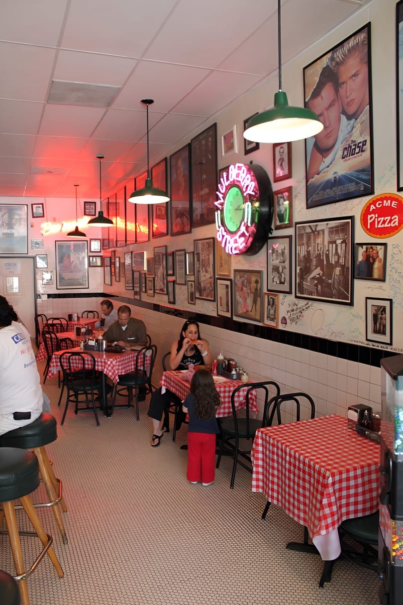 Diners seated at red and white checkered tables in a restaurant with movie posters and neon signs.