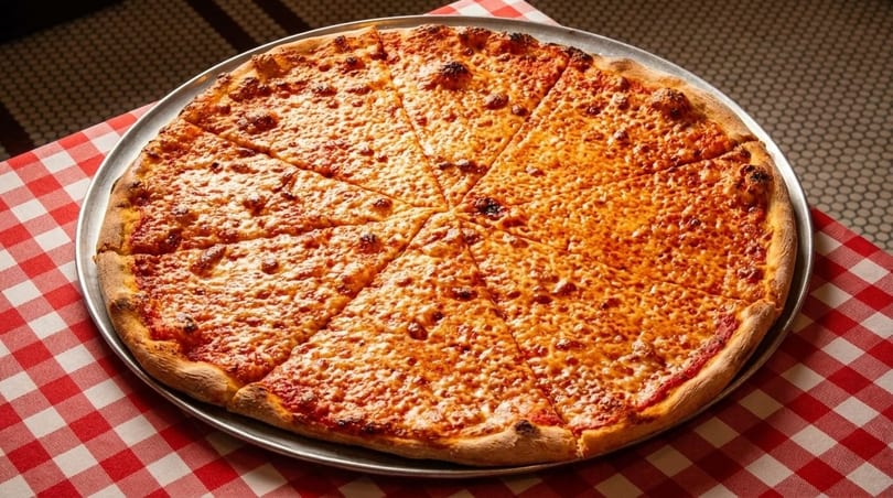 A large, sliced cheese pizza on a metal tray, resting on a red and white checkered tablecloth.