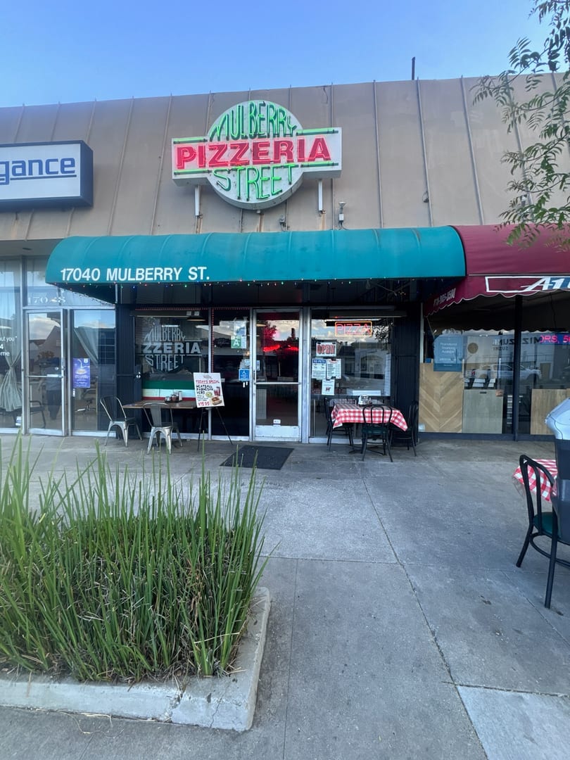 Mulberry Pizzeria Street storefront with a green awning, outdoor seating, and a neon sign.