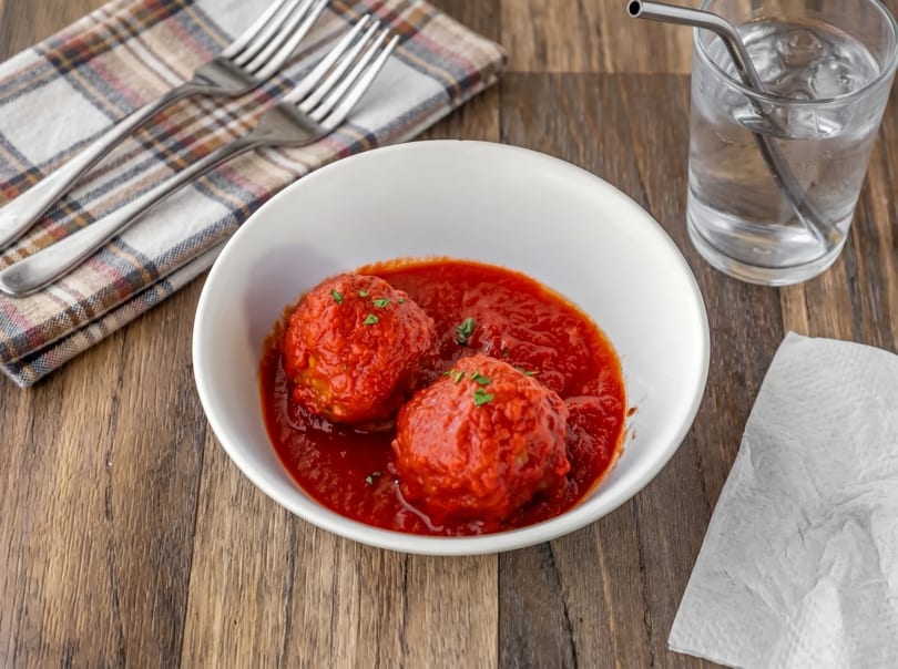 Two large meatballs in red sauce, garnished with parsley, in a white bowl on a wooden table.